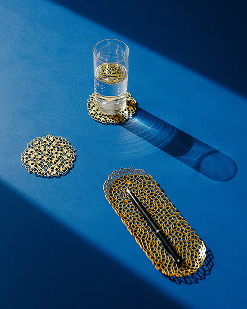 Penrose coasters and pen tray in brass on a blue surface beside a glass