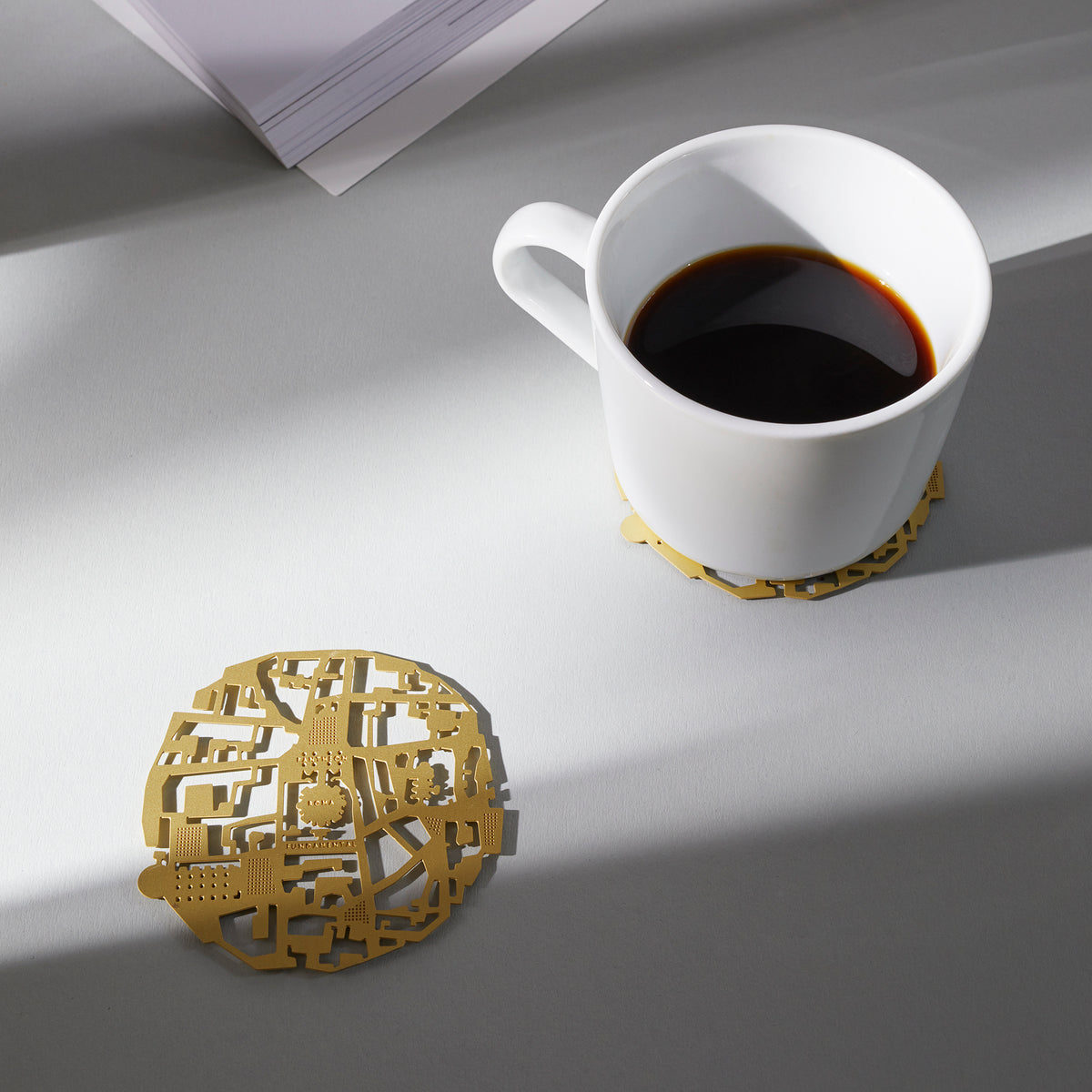 A Roma brass coaster under a cup of coffee, its pair alongside